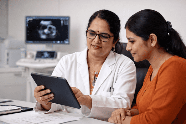 Indian gynecologist Dr. Asha Sharma reviewing medical reports on a tablet with a female patient in a modern gynecology clinic, ultrasound screen visible in the background.
